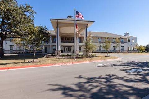 Front exterior view of a two-story senior living facility building with a covered entrance supported by columns, two flagpoles with the American and Texas flags, a circular driveway, and trees on a clear sunny day.