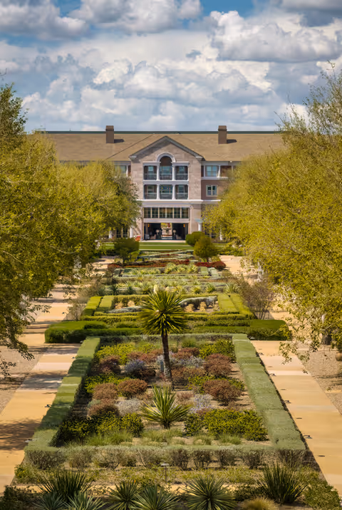 A landscaped garden with neatly trimmed hedges and various plants leading to a large multi-story building under a partly cloudy sky.