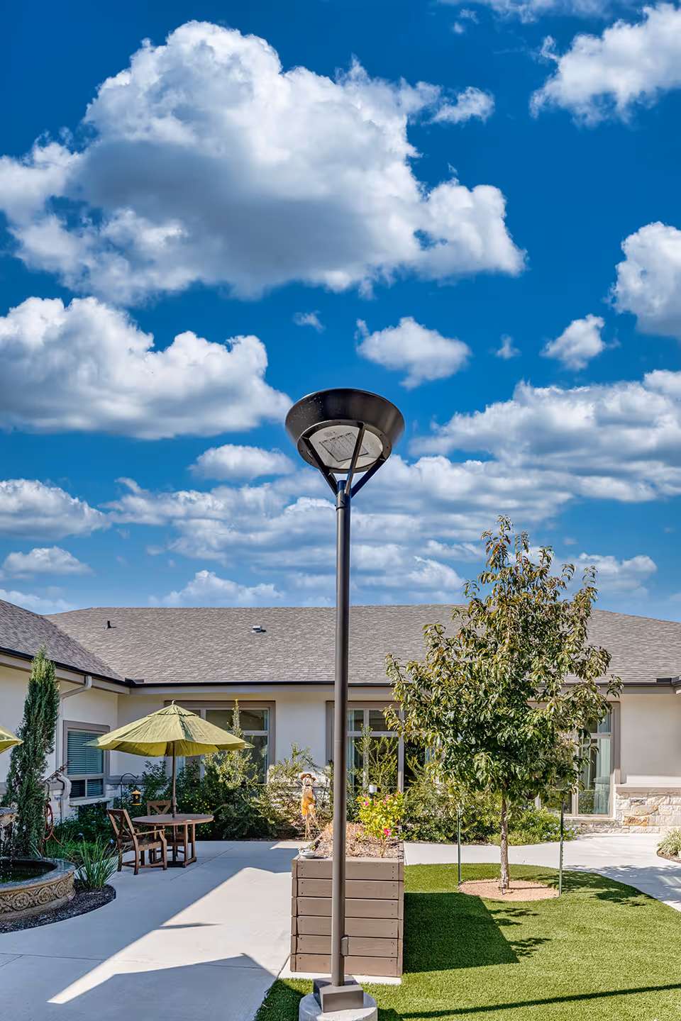 Outdoor courtyard area at Volante Senior Living of Sage Spring featuring a tall lamp post in the center, a small tree, green grass, a patio with a table and chairs under a green umbrella, and a building with windows in the background under a blue sky with scattered clouds.