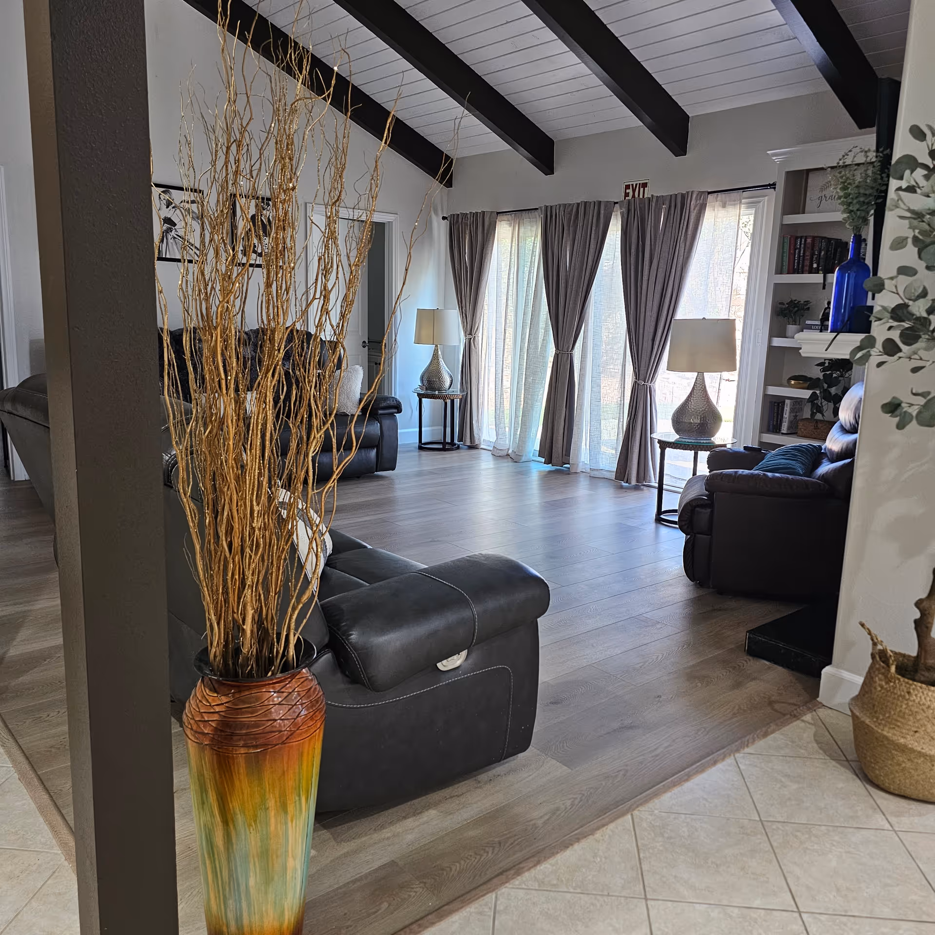 A bright living room with light wood flooring and a high ceiling with exposed dark beams. The room features black leather sofas, two side tables with lamps, large windows covered with sheer curtains, and a decorative tall vase with dried branches in the foreground. There is a built-in bookshelf with books and decorative items on the right side.
