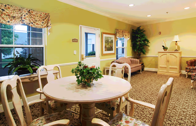 Communal senior living dining area with a round table and chairs, upholstered sofas, plants, and windows.