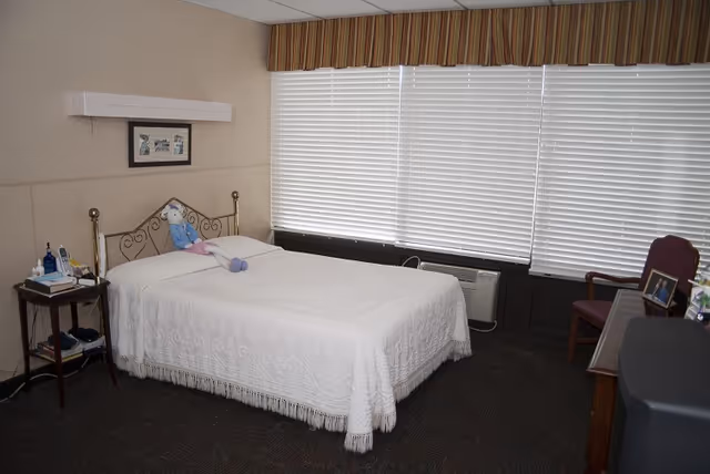 A simple bedroom with a double bed covered in a white bedspread, a bedside table, large windows with closed blinds, and a chair and desk.