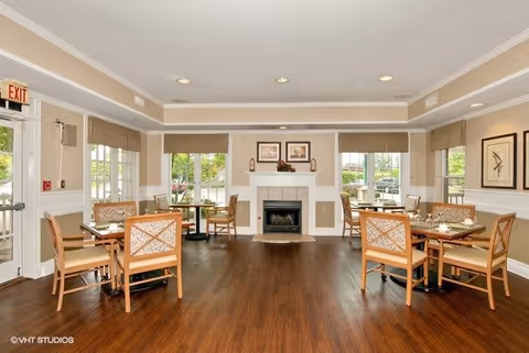 A bright dining room with wooden floors and several tables surrounded by chairs. The room features large windows with beige blinds, a white fireplace with framed artwork above it, and neutral-colored walls. There is an exit door on the left side and decorative wall art on the right.