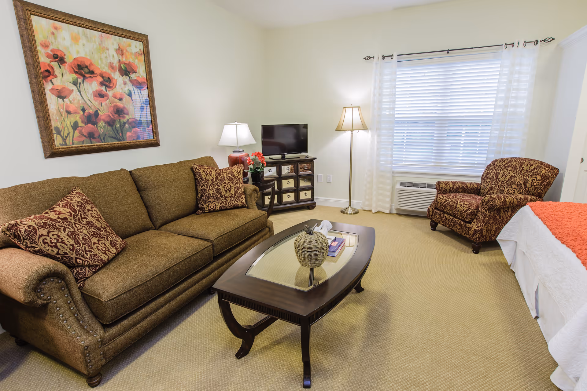 A cozy living area with a brown sofa adorned with patterned cushions, a matching armchair, a wooden coffee table with a decorative basket and books, a small TV on a cabinet, a floor lamp, and a window with white blinds and sheer curtains. A bed with white and orange bedding is partially visible on the right side of the image. A framed floral painting hangs on the wall above the sofa.