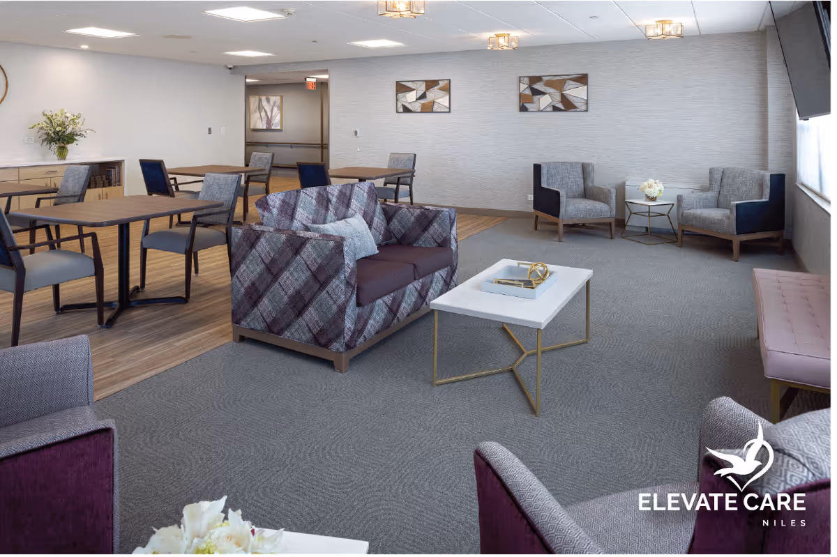 A spacious and well-lit common area in a senior living facility featuring a mix of seating options including patterned armchairs, a loveseat, and dining tables with chairs. The room has a carpeted floor with a section of wood flooring near the dining tables, modern wall art, and a large window allowing natural light. A white coffee table with decorative items is in the center, and a sideboard with a flower arrangement is visible in the background.