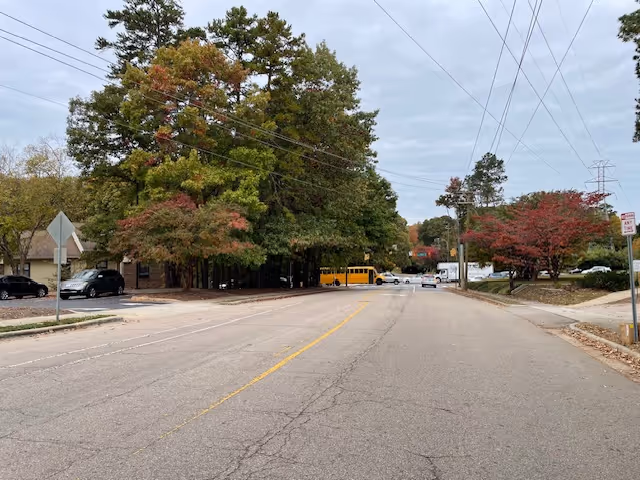 Wide residential street lined with trees and parked cars, a yellow school bus visible in the distance under an overcast sky.