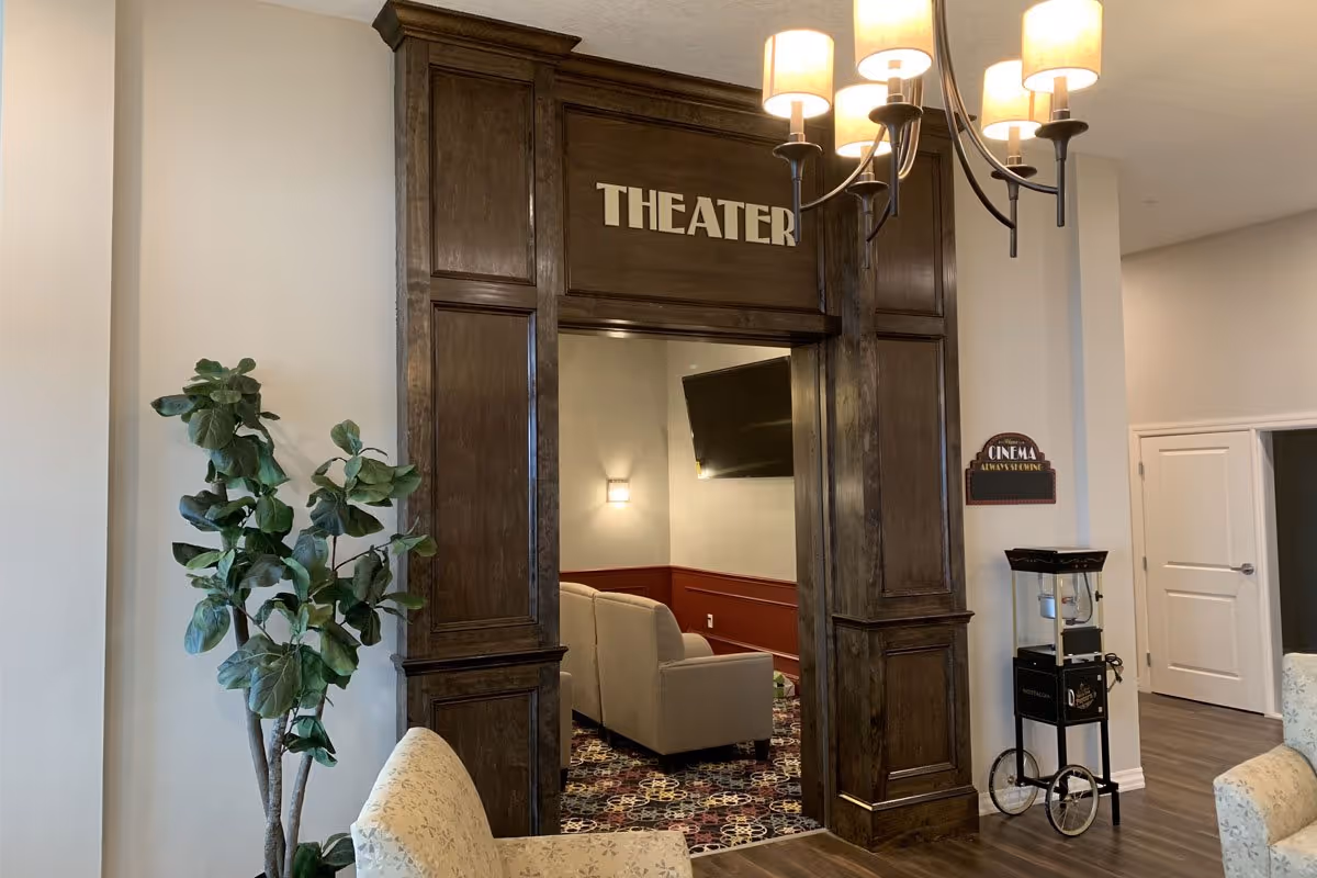 Entrance to a theater room in an assisted living facility with dark wood paneling around the doorway labeled 'THEATER'. Inside, there are beige sofas, a wall-mounted TV, and a patterned carpet. A popcorn machine is positioned near the entrance, and a chandelier with multiple lamps hangs from the ceiling. A potted plant and upholstered chairs are also visible in the foreground.