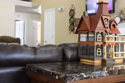 A close-up view of a dark marble tabletop with a detailed miniature wooden house model on it. Behind the table is a dark brown leather couch, and in the background, there is a beige wall with a white door, a wall-mounted light fixture, and a wall decoration. A television screen is partially visible above the miniature house.