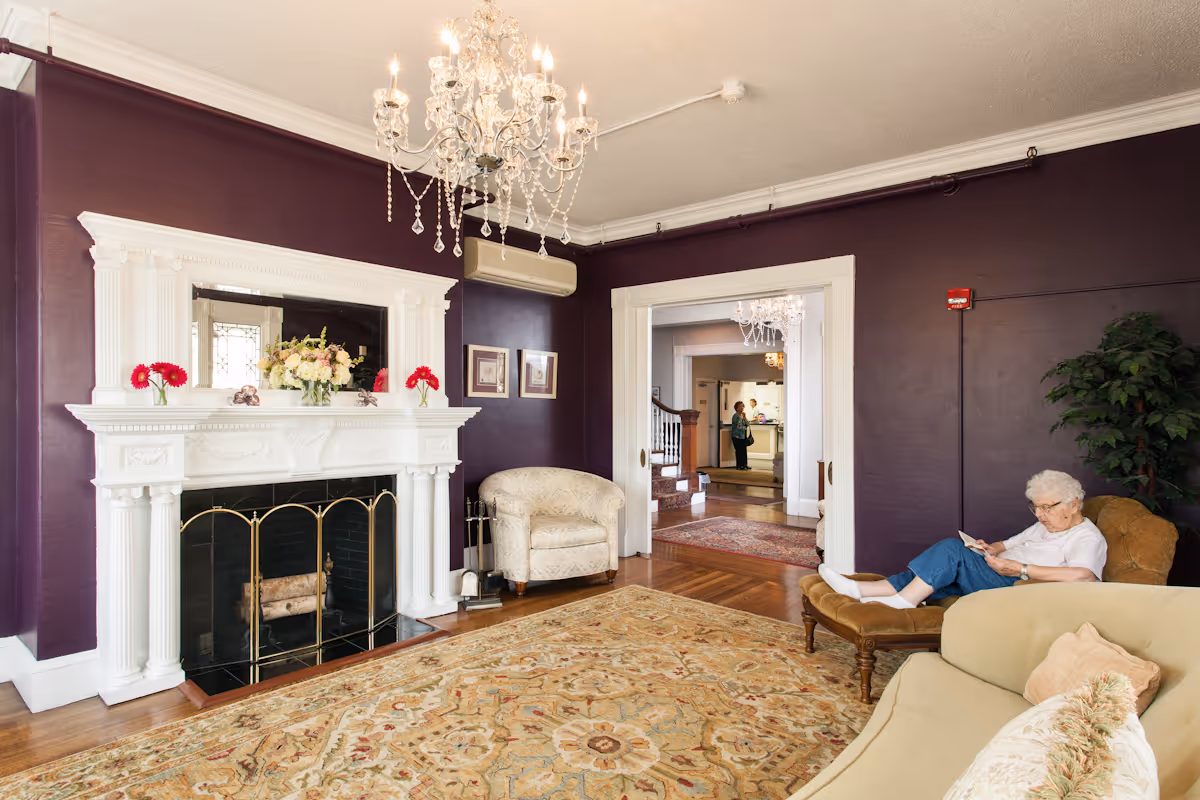 An elderly woman reads on a chaise in a cozy living room with a white fireplace, chandelier, and purple walls.