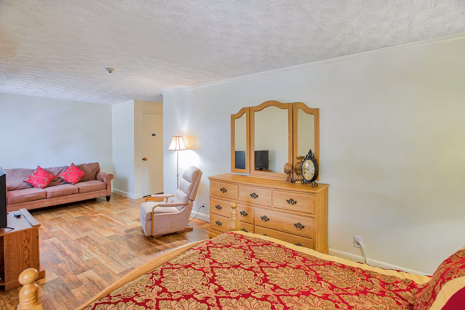A senior living facility room featuring a bed with a red and gold patterned bedspread in the foreground, a wooden dresser with a three-panel mirror and decorative items including a clock, a beige armchair, a floor lamp, a brown sofa with two red pillows, and a TV on a wooden stand. The room has wooden flooring and light-colored walls.