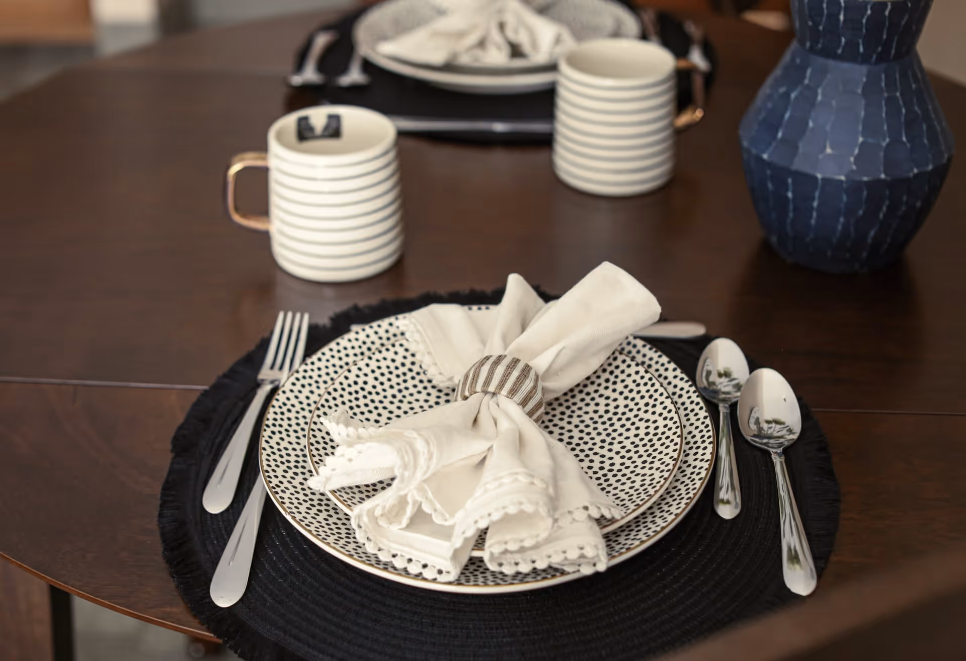Close-up view of a dining table set with a black placemat, two patterned plates, a white cloth napkin with a striped napkin ring, silverware including a fork, knife, and two spoons, two striped mugs, and a blue vase.