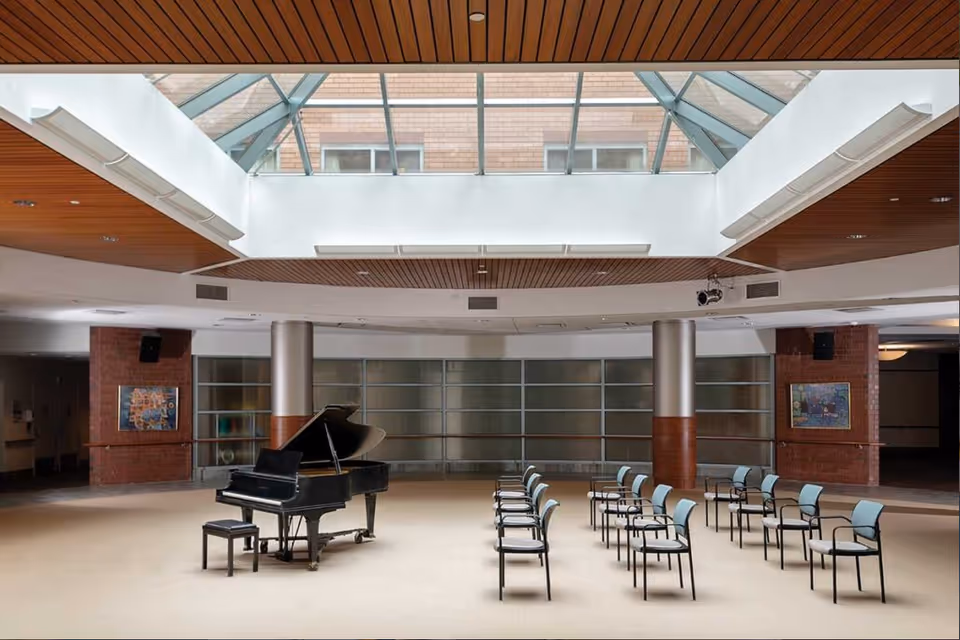 A spacious indoor common area with a grand piano and a matching piano bench in the center. There are three rows of chairs arranged facing the piano. The ceiling features a large skylight allowing natural light to illuminate the room. The walls have a combination of brick and glass panels, and there are two framed artworks on the brick walls.