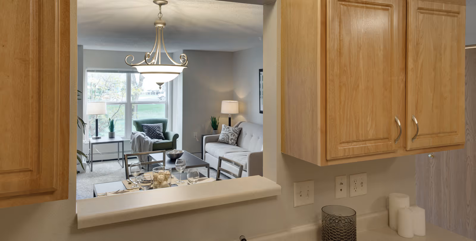 View from a kitchen pass-through window into a living room area with a green armchair, a gray sofa with decorative pillows, a coffee table, and a dining table set with glassware and a centerpiece. The kitchen has wooden cabinets, a countertop with candles, and a decorative container.