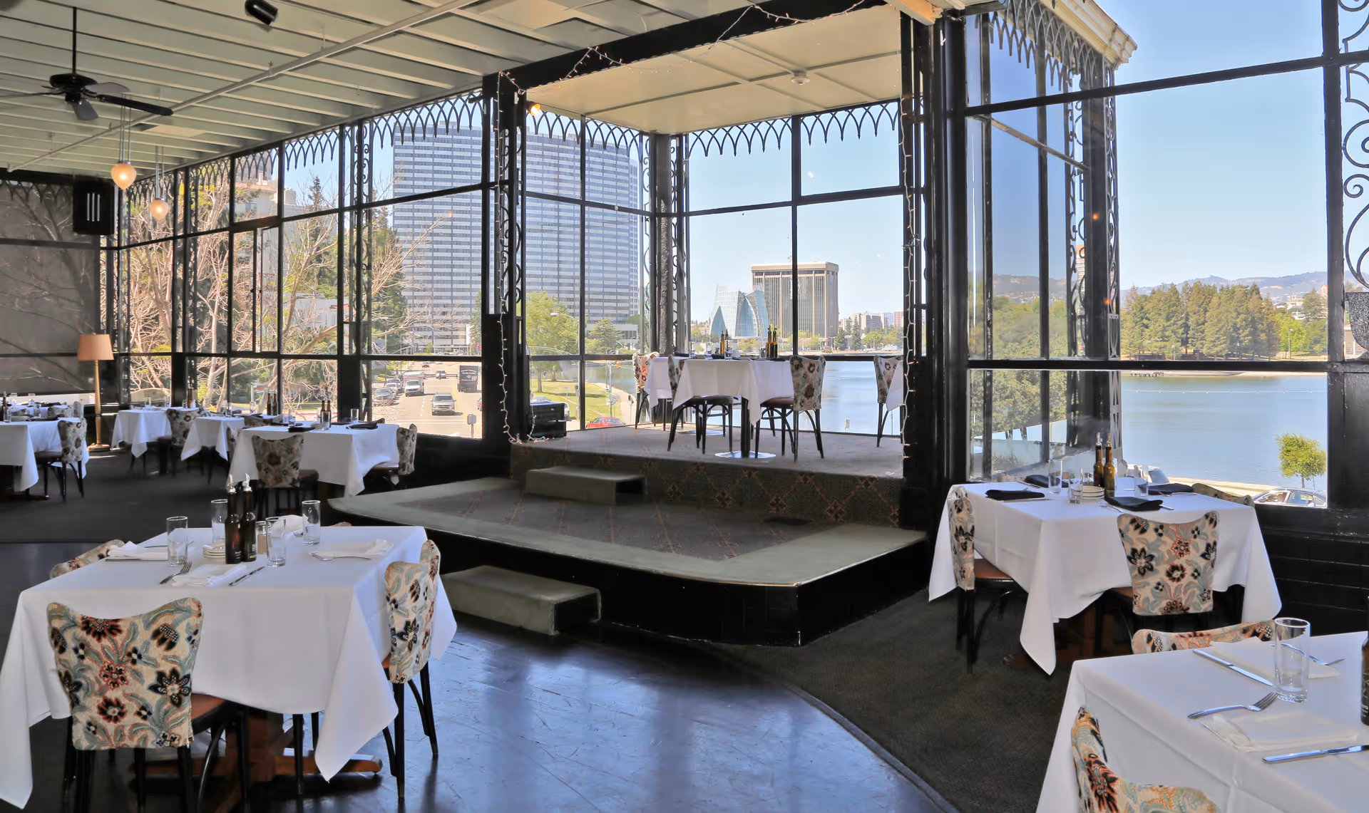 Dining room with white-tablecloth tables and floral chairs overlooking a lake through large floor-to-ceiling windows.