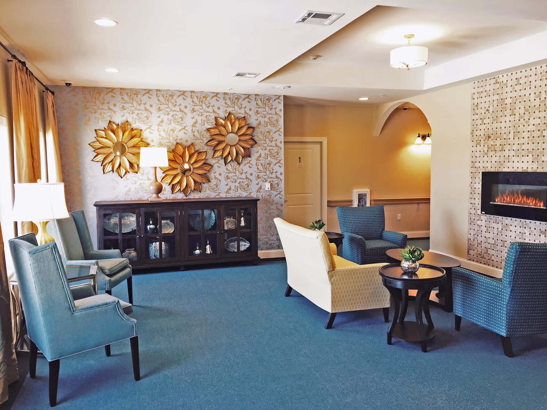 A cozy sitting area in a memory care facility with blue carpet, a cream-colored loveseat, three blue armchairs, two round wooden side tables with small plants, a modern electric fireplace set in a tiled wall, and a dark wooden cabinet against a wallpapered wall with three decorative sunburst mirrors and a table lamp.