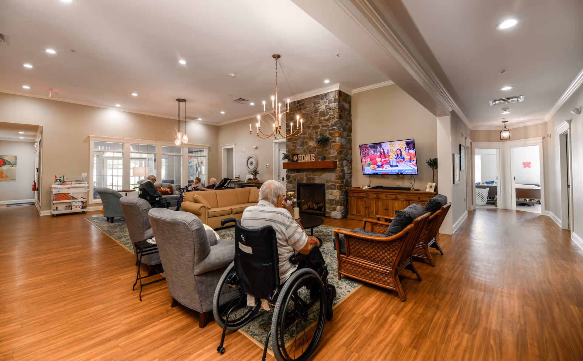 A spacious senior living common area with wooden flooring, a stone fireplace with a 'HOME' sign on the mantle, and a flat-screen TV mounted on the wall. Several seniors are seated on sofas and chairs, including one person in a wheelchair facing the TV. The room is well-lit with ceiling lights and chandeliers, and there is a hallway leading to other rooms visible on the right side.