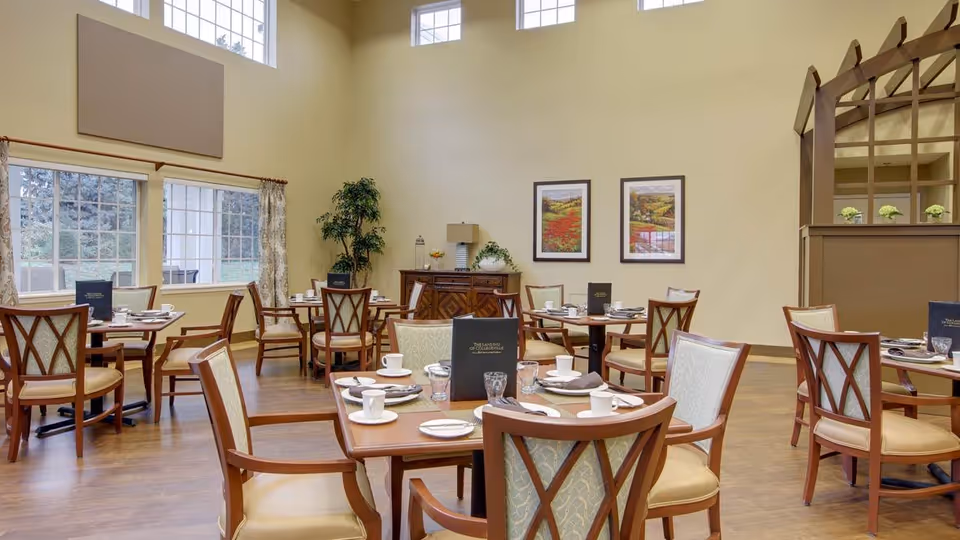 A bright dining room with several wooden tables and chairs arranged neatly. Each table is set with white cups, plates, glasses, and menus. The room has large windows letting in natural light, beige walls, two framed landscape paintings, and a decorative wooden structure on the right side. There is also a sideboard with a lamp and plants.