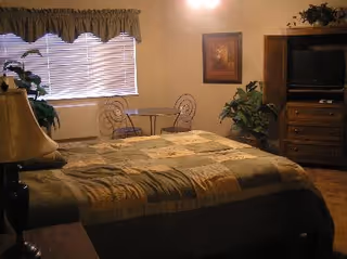 Cozy bedroom with a bed covered in a patchwork quilt, a bedside lamp, a small round table with two chairs by a window, a TV armoire, and plants.