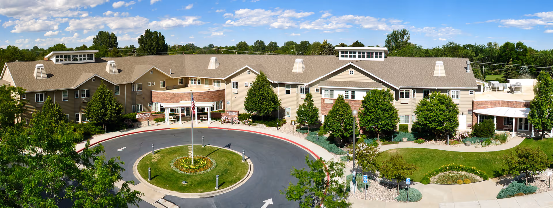 Panoramic front view of a large two-story senior living facility with a circular driveway, flagpole, and landscaped grounds.