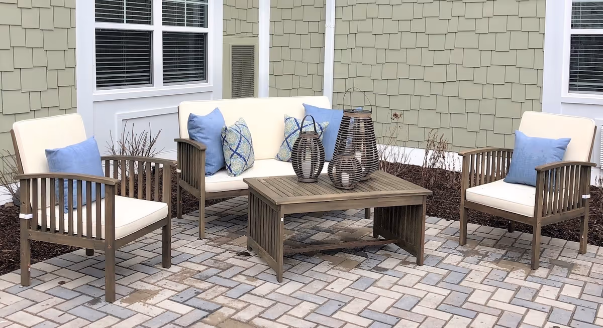 Outdoor patio area with a wooden seating set including two armchairs and a loveseat with white cushions and blue decorative pillows. A wooden coffee table holds three decorative lanterns. The patio is paved with light-colored bricks and is adjacent to a building with light green siding and white trim around windows.