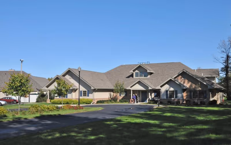 Exterior view of a single-story senior living community building with a driveway, green lawn, small trees, and a clear blue sky. Two people are standing near the entrance of the building.