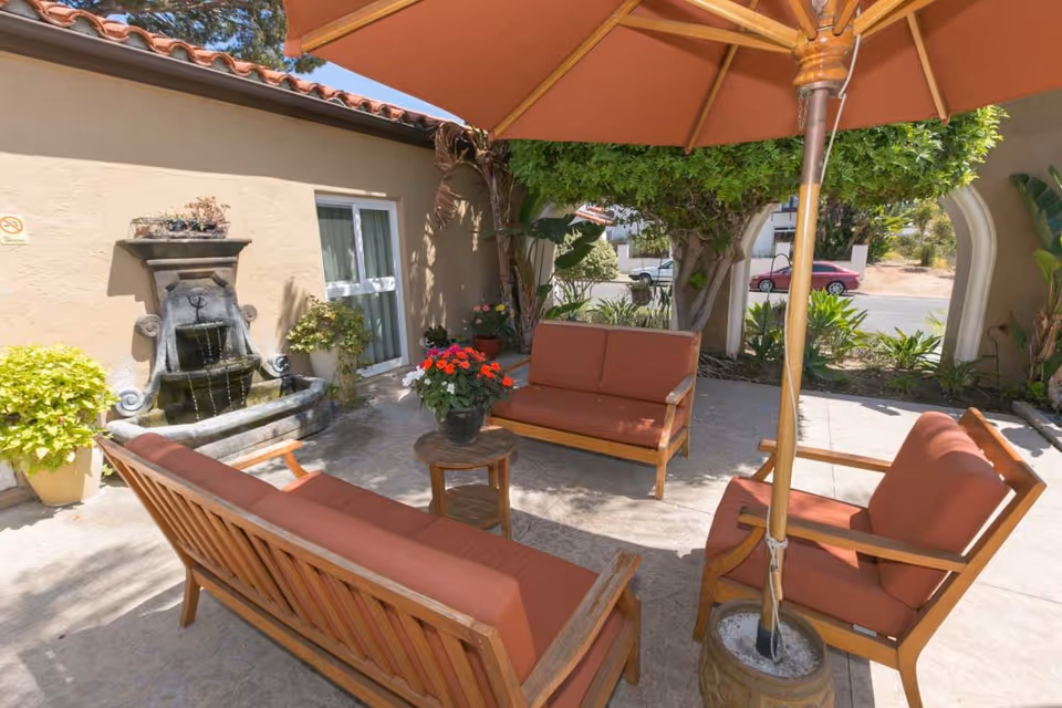 Outdoor patio area with wooden seating furniture featuring rust-colored cushions arranged around a small round table with a flower pot. A large umbrella provides shade. There is a decorative stone water fountain against a beige wall, potted plants, and greenery including a tree and shrubs. A street and parked cars are visible in the background.