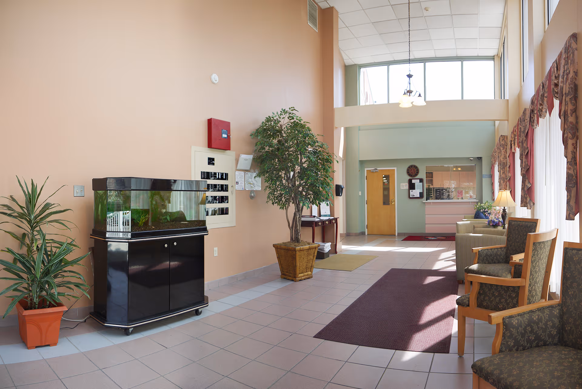 A bright and spacious lobby area with large windows letting in natural light. The room features a fish tank on a black stand, several potted plants, a maroon rug, and multiple chairs with wooden arms and patterned upholstery. There is a reception desk in the background with a clock and bulletin board on the wall behind it. The walls are painted in soft pastel colors.