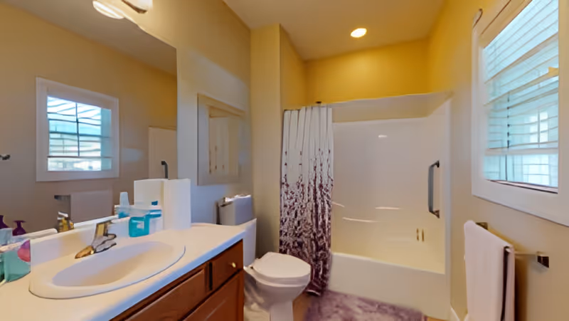Bright bathroom with a sink and vanity, toilet, bathtub with a shower curtain, window, and towel rack.
