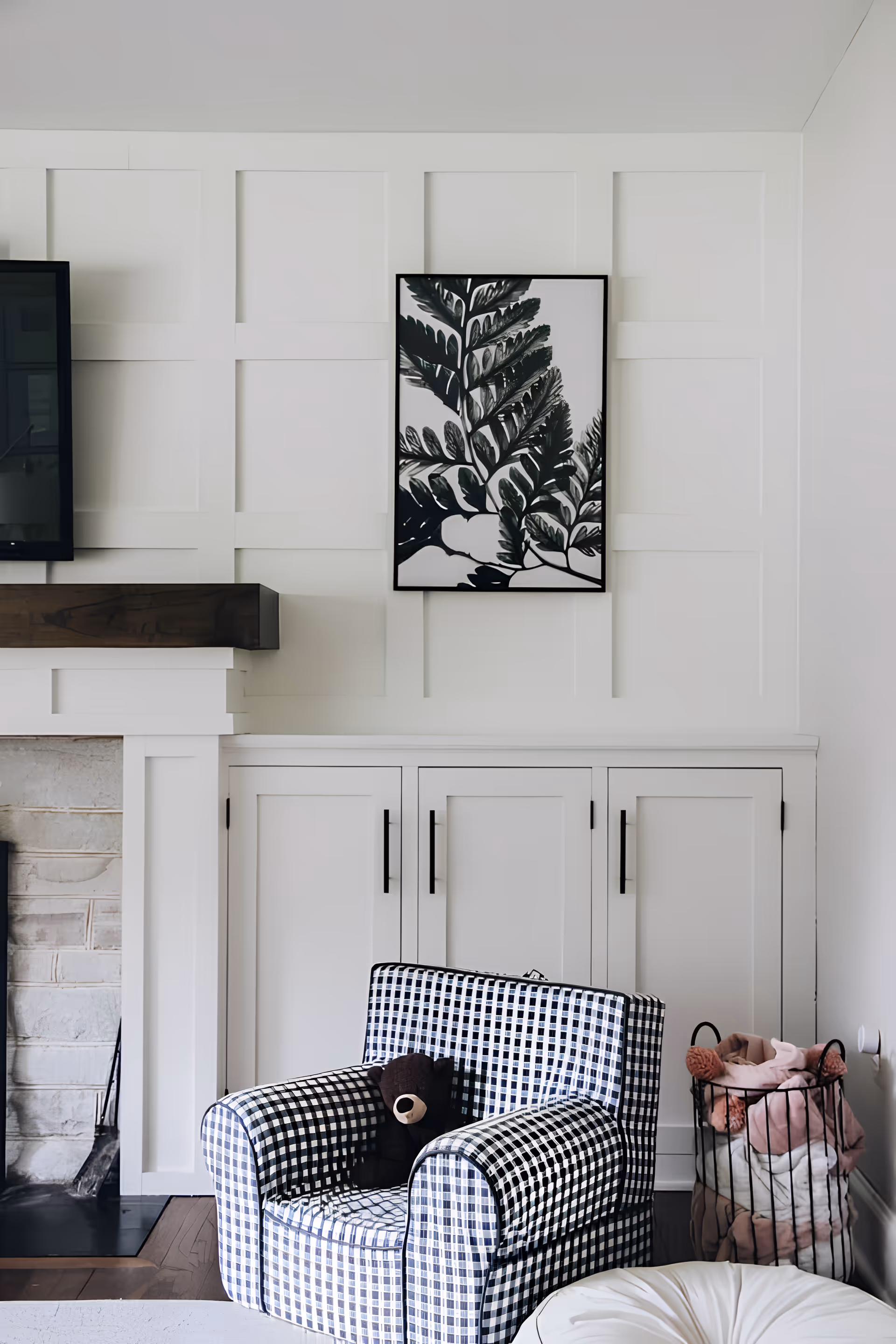 A cozy living room corner featuring a black and white checkered armchair with a small brown teddy bear on it, a white paneled wall with a framed black and white leaf artwork, white cabinetry with black handles, a fireplace with a wooden mantel, and a wire basket filled with pink and white blankets.