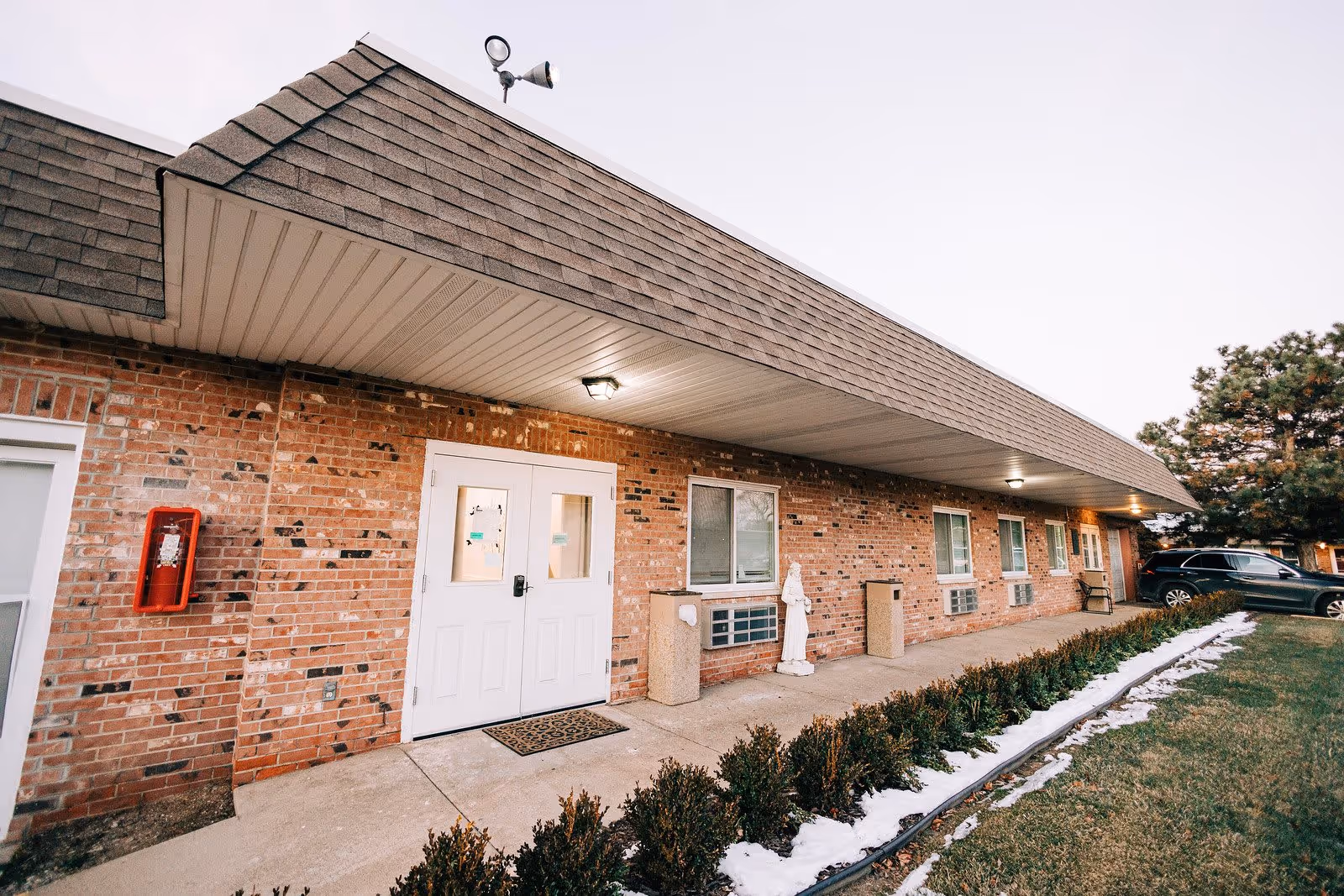 Exterior view of a single-story brick building with a shingled roof, white double doors, several windows, outdoor lights, a statue, and a parked car on the right side. There is a small garden bed with shrubs and patches of snow along the sidewalk in front of the building.