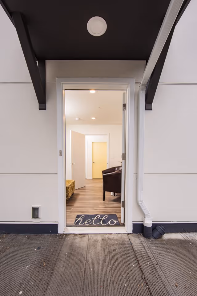View through an open exterior door into a room with wooden flooring, a brown armchair, and a partially visible yellow door in the background. A doormat with the word 'hello' is placed at the entrance.