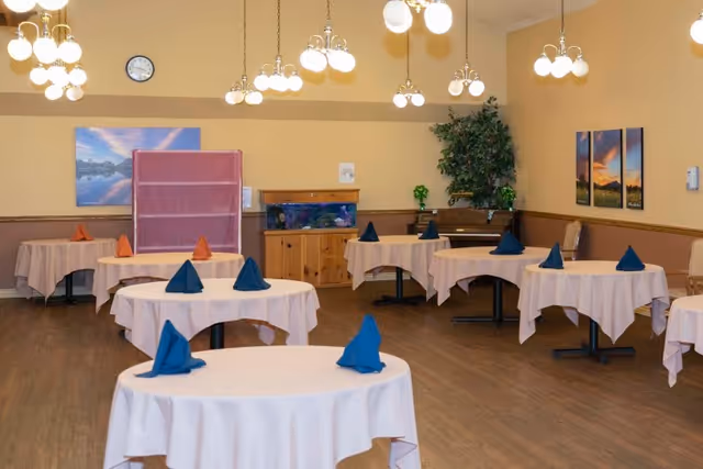 A dining room with round tables covered in white tablecloths, each set with folded cloth napkins in blue and orange colors. The room has wooden flooring, beige walls, multiple hanging light fixtures with globe bulbs, a clock on the wall, framed landscape pictures, a fish tank on a wooden cabinet, a piano, and a large potted plant in the corner.