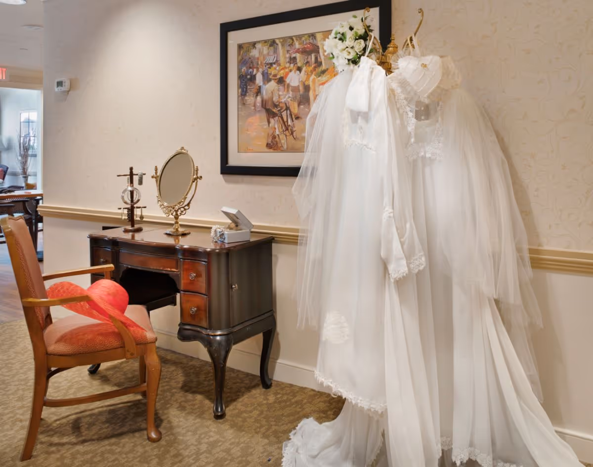 A vintage-style vanity table with a mirror and jewelry stand, accompanied by a wooden chair with a red cushion. Next to the vanity, two white wedding dresses with veils and a floral headpiece are displayed on a coat rack against a wallpapered wall. A framed painting depicting a street scene hangs above the dresses.