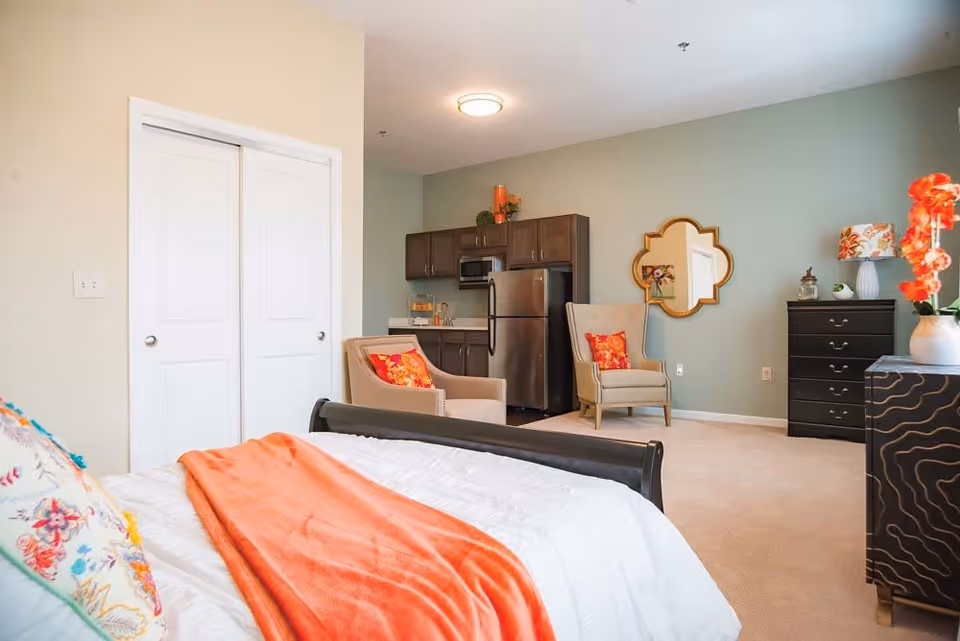 A bright and cozy bedroom featuring a bed with white bedding and an orange throw blanket, two beige armchairs with orange floral pillows, a small kitchenette with dark cabinets and stainless steel appliances, a decorative mirror on a green accent wall, a black dresser, and a vase with orange flowers on a side table.