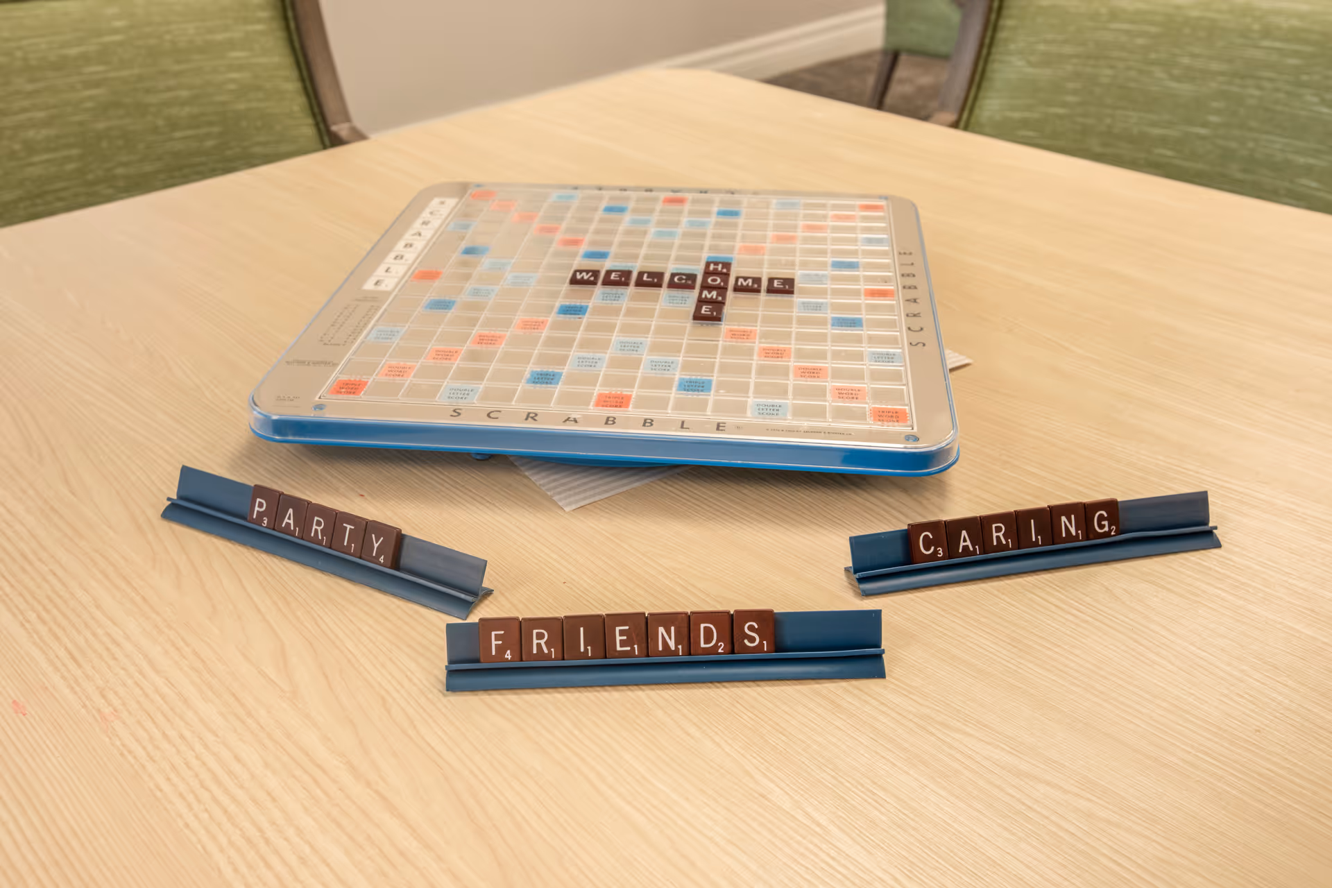 A Scrabble board on a light wooden table with the word 'WELCOME HOME' spelled out on the board. In front of the board are three Scrabble tile racks with the words 'PARTY', 'FRIENDS', and 'CARING' spelled out. Two green chairs are partially visible in the background.