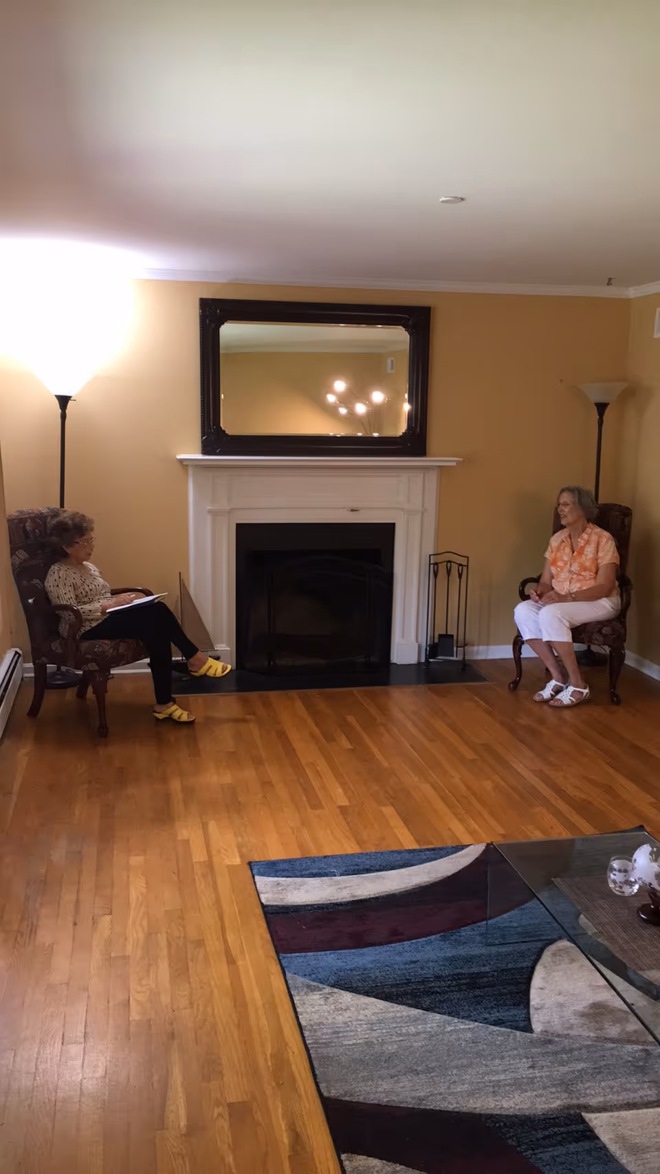 Two elderly women sitting in armchairs facing each other in a living room with wooden floors, a fireplace with a large mirror above it, two floor lamps, and a colorful area rug partially visible in the foreground.