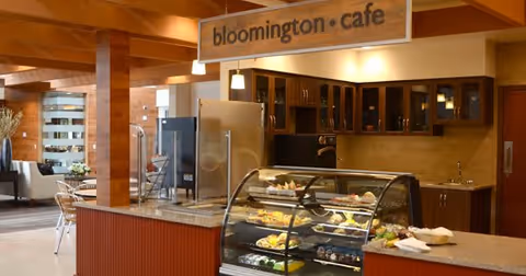 Interior view of a cafe area named Bloomington Cafe with a glass display case filled with pastries and desserts, wooden cabinetry, a refrigerator, and seating area with tables and chairs in the background.