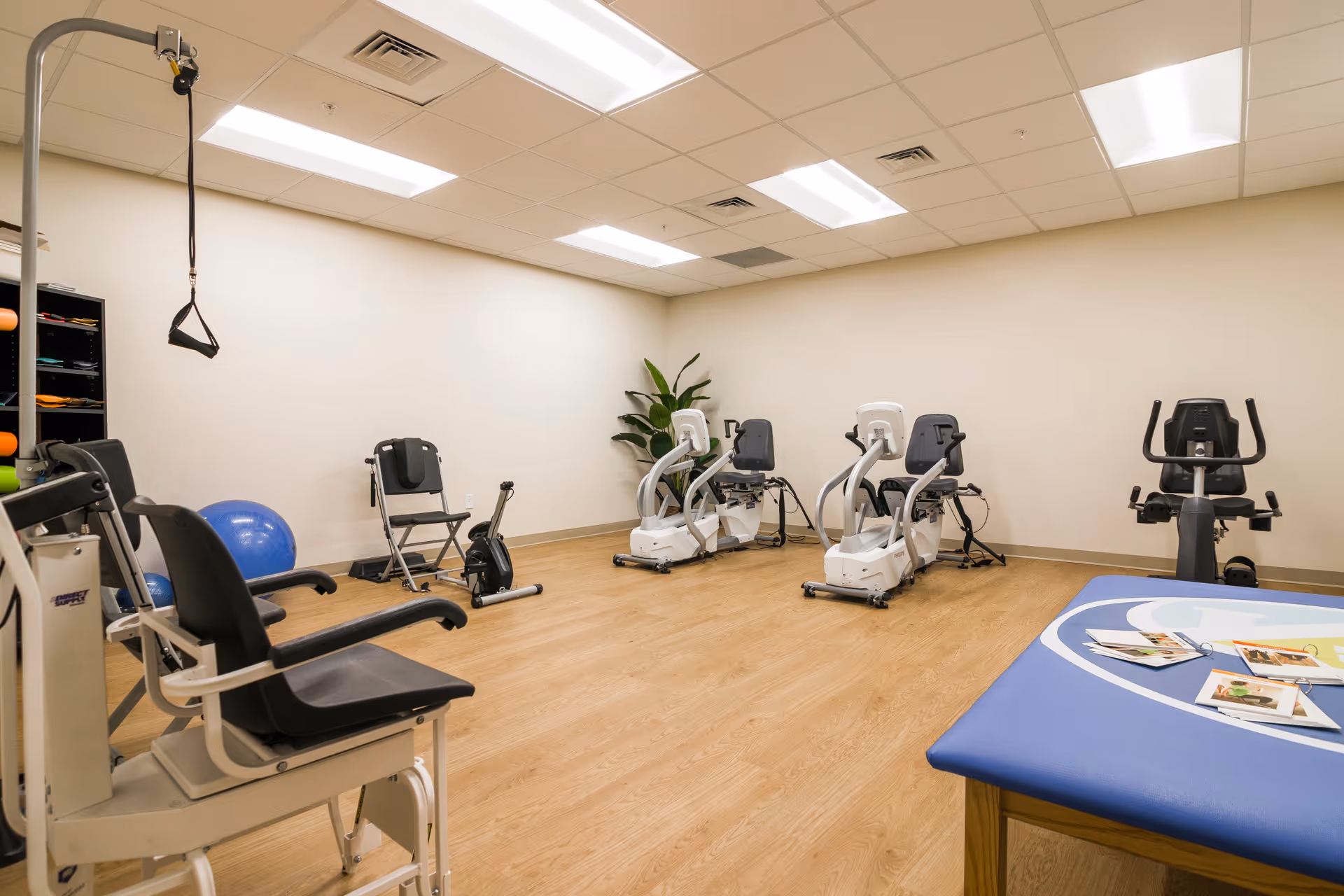 A bright exercise/therapy room with multiple recumbent exercise bikes, fitness equipment, and a treatment table.