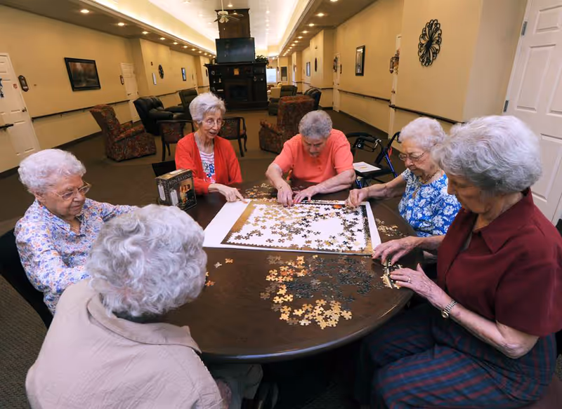 Five elderly women sitting around a round table in a common area, working together on assembling a large jigsaw puzzle. The room has beige walls, framed pictures, and several armchairs in the background.