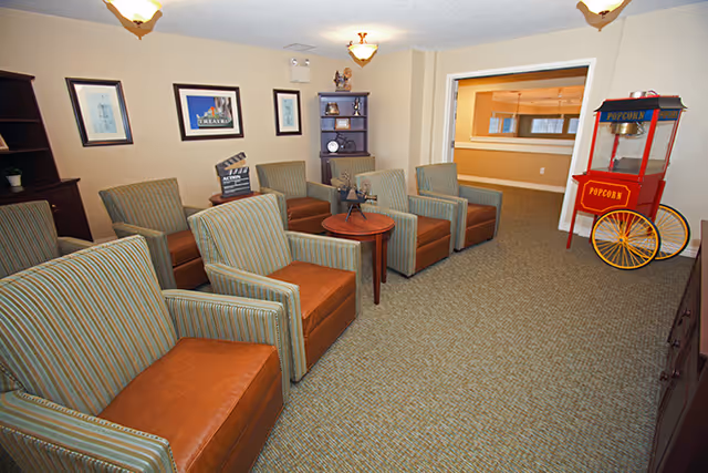 A cozy common area with six striped armchairs arranged in two rows facing a small round wooden table. The room has beige walls adorned with framed pictures and a small dark wooden shelf holding decorative items. In the corner, there is a vintage-style red popcorn machine on wheels. The carpet is patterned in neutral tones, and ceiling lights illuminate the space.