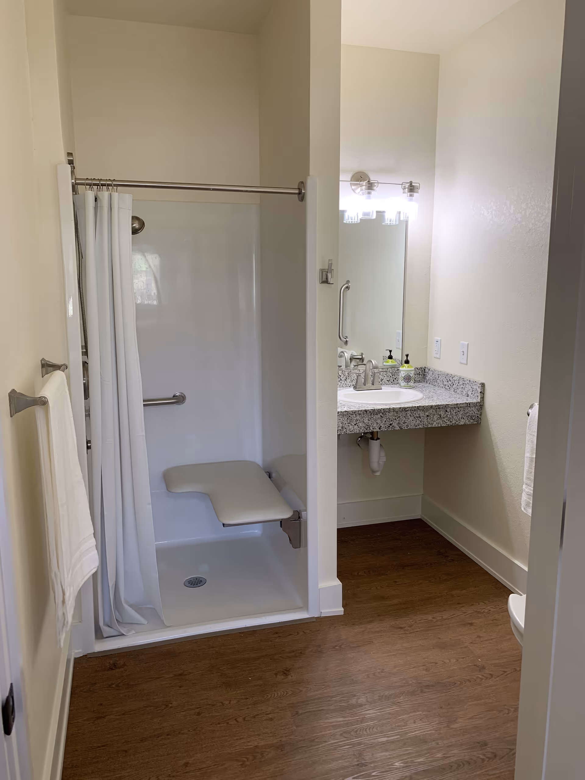 A bathroom with a walk-in shower featuring a fold-down seat and grab bars, a white shower curtain, a granite countertop with a sink, a mirror above the sink, and a wall-mounted light fixture. The floor has wood-like flooring and there are white towels hanging on towel racks.
