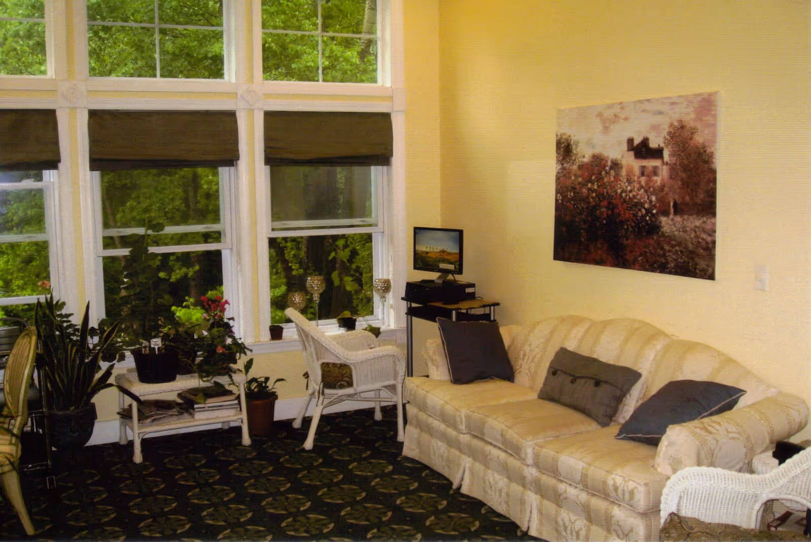 A cozy living room with a cream-colored patterned sofa adorned with three dark pillows. Next to the sofa is a white wicker chair and a small table with several potted plants. Large windows with brown blinds reveal green trees outside. A small computer setup is on a stand near the sofa, and a painting of a house with flowers hangs on the yellow wall.