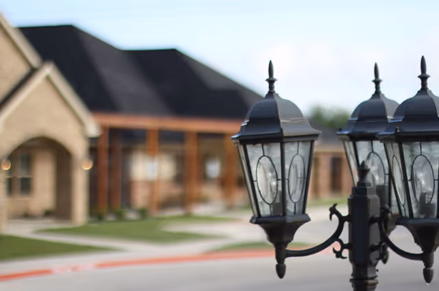 Decorative black wrought-iron lamppost in focus with a blurred single-story brick building and driveway in the background.