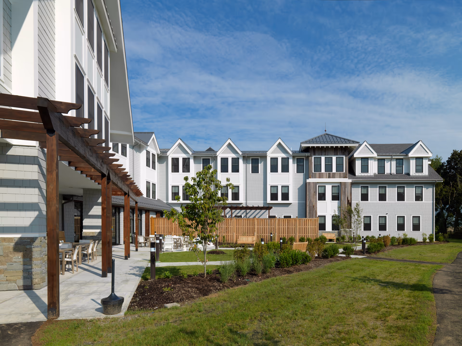 Exterior front view of a three-story senior living building with a pergola-covered patio, landscaped lawn, and blue sky.