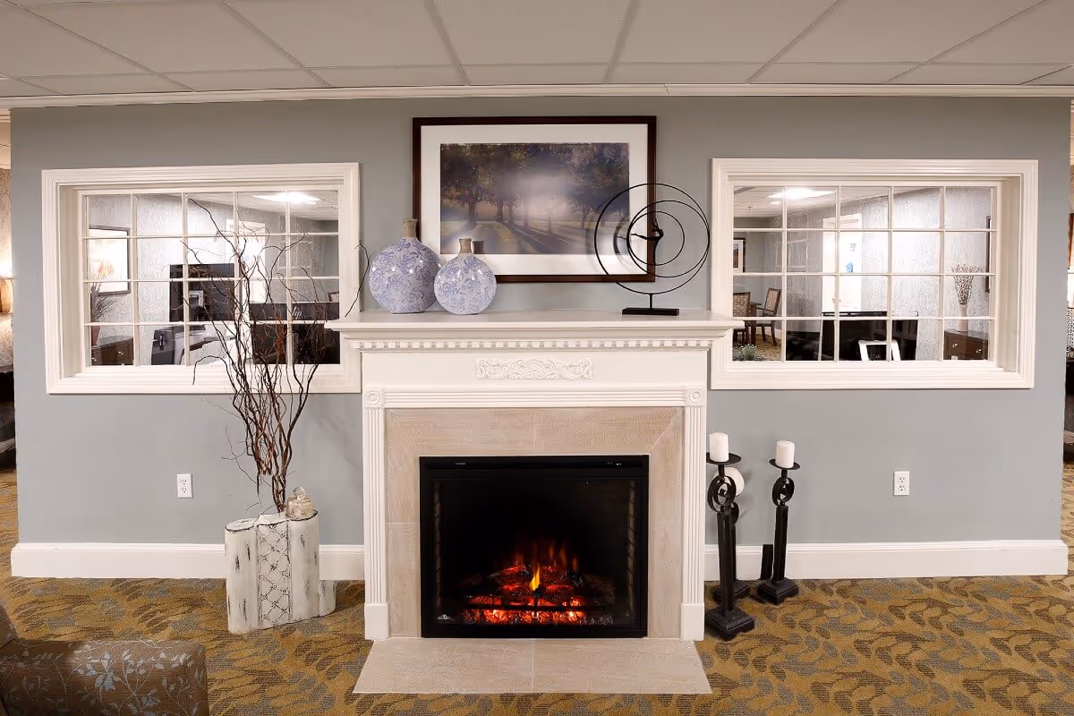 A cozy indoor fireplace with a white mantel decorated with two blue patterned vases and a modern circular metal sculpture. Above the mantel is a framed picture of a tree-lined path. On either side of the fireplace are two large windows with white frames showing adjacent rooms. The floor has a patterned carpet, and there are two black candle holders with white candles on the right side of the fireplace and a tall vase with decorative branches on the left.
