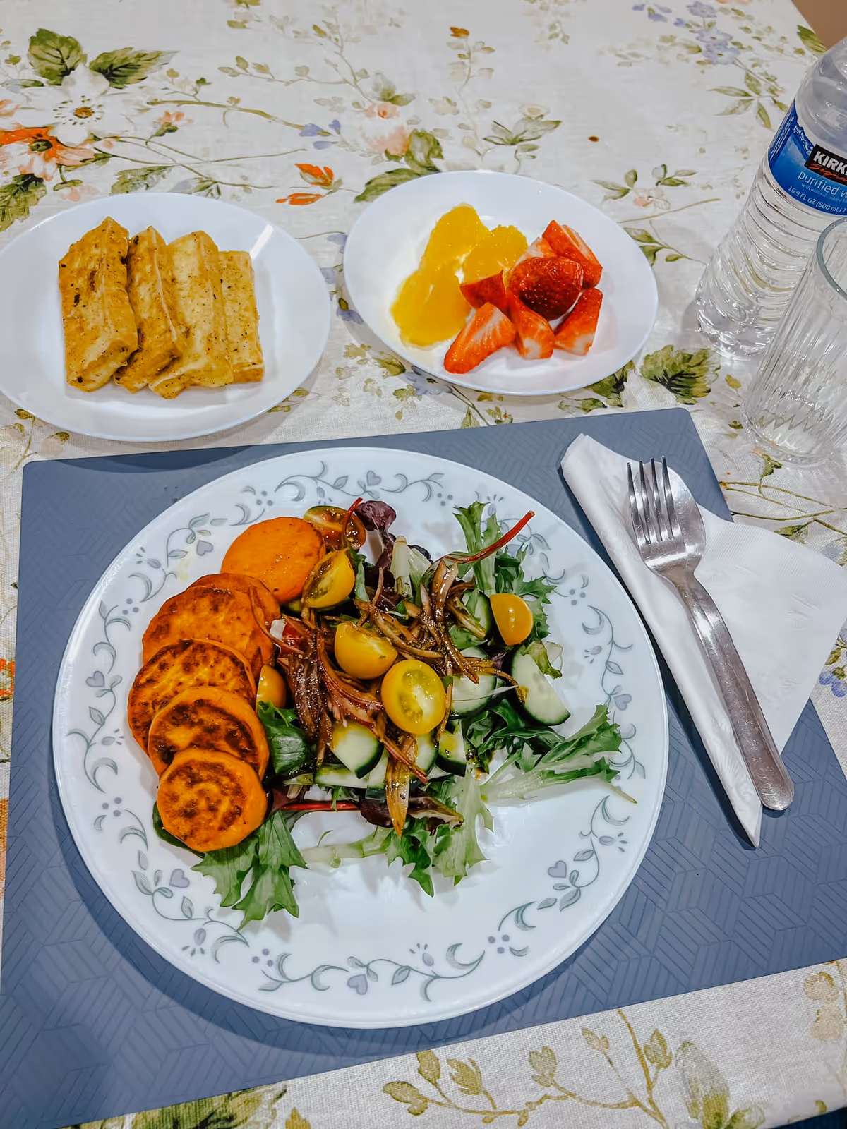 A meal set on a floral tablecloth with a plate containing a salad of mixed greens, yellow cherry tomatoes, cucumber slices, and small fried fish, accompanied by slices of cooked sweet potato. There is a small bowl with grilled chicken strips and another bowl with orange slices and strawberry halves. A bottle of Kirkland purified water, an empty glass, and a fork and spoon wrapped in a white napkin are also visible.
