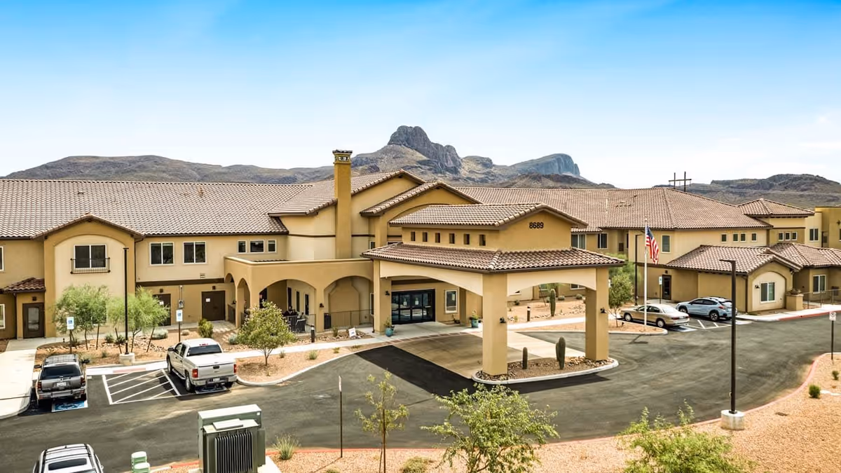 Exterior view of a large senior living facility building with a covered entrance and multiple windows. The building has a beige stucco finish with a tiled roof. There are several parked cars in the parking lot, desert landscaping with small trees and cacti, and mountains visible in the background under a clear blue sky.