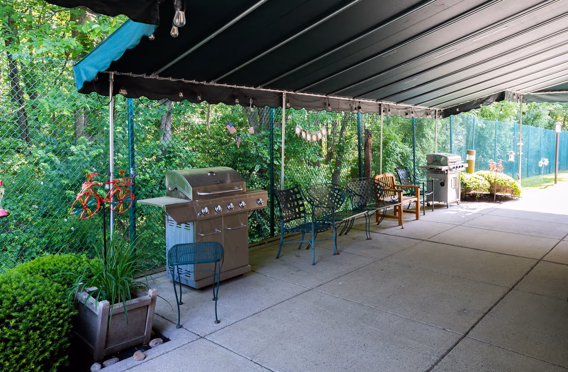 Covered outdoor patio area with two stainless steel grills, several metal and wooden chairs, a small metal table, and decorative plants. The area is enclosed by a green chain-link fence with trees and greenery visible beyond it.