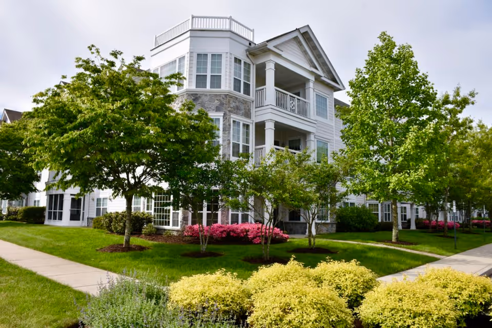 White three-story senior living building with balconies and large windows set among trees and landscaped lawns.