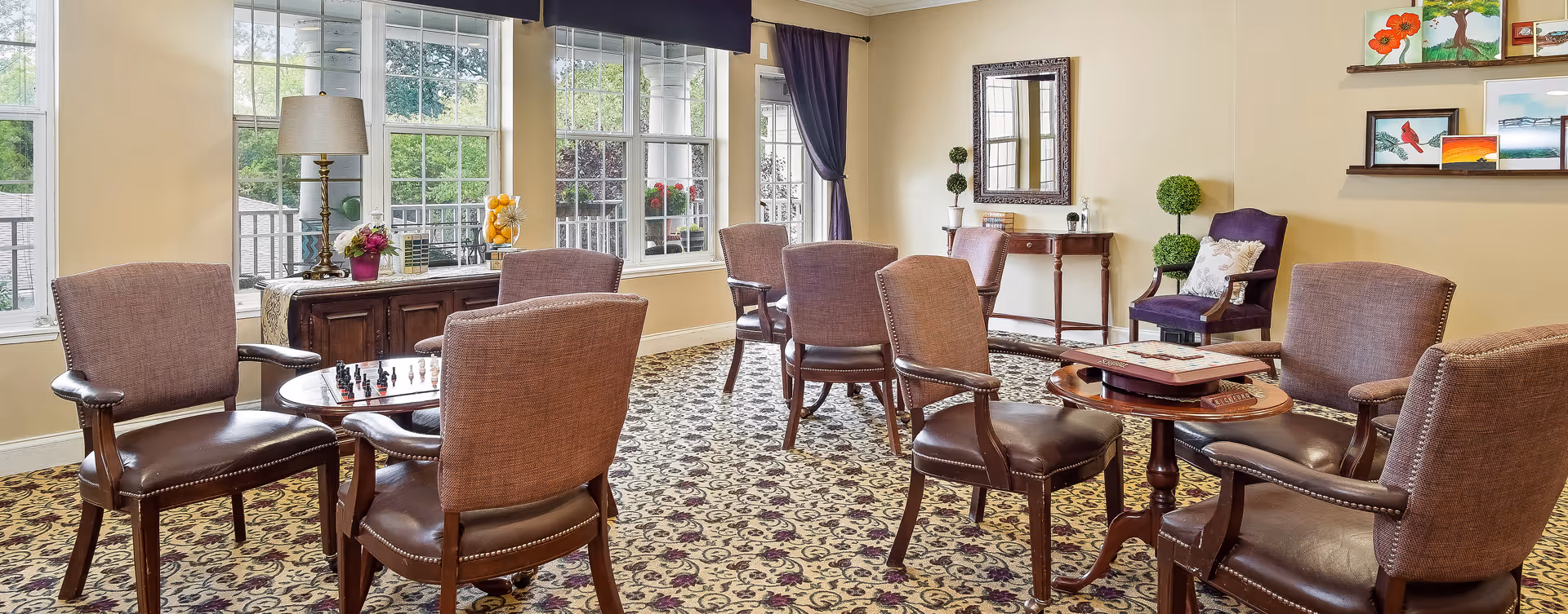 A bright and inviting common area with multiple upholstered chairs arranged around small wooden tables. One table has a chess set, and another has a Scrabble game. Large windows let in natural light and offer views of greenery outside. The room features patterned carpet, a sideboard with a lamp and flowers, a mirror on the wall, and shelves with colorful artwork and plants.